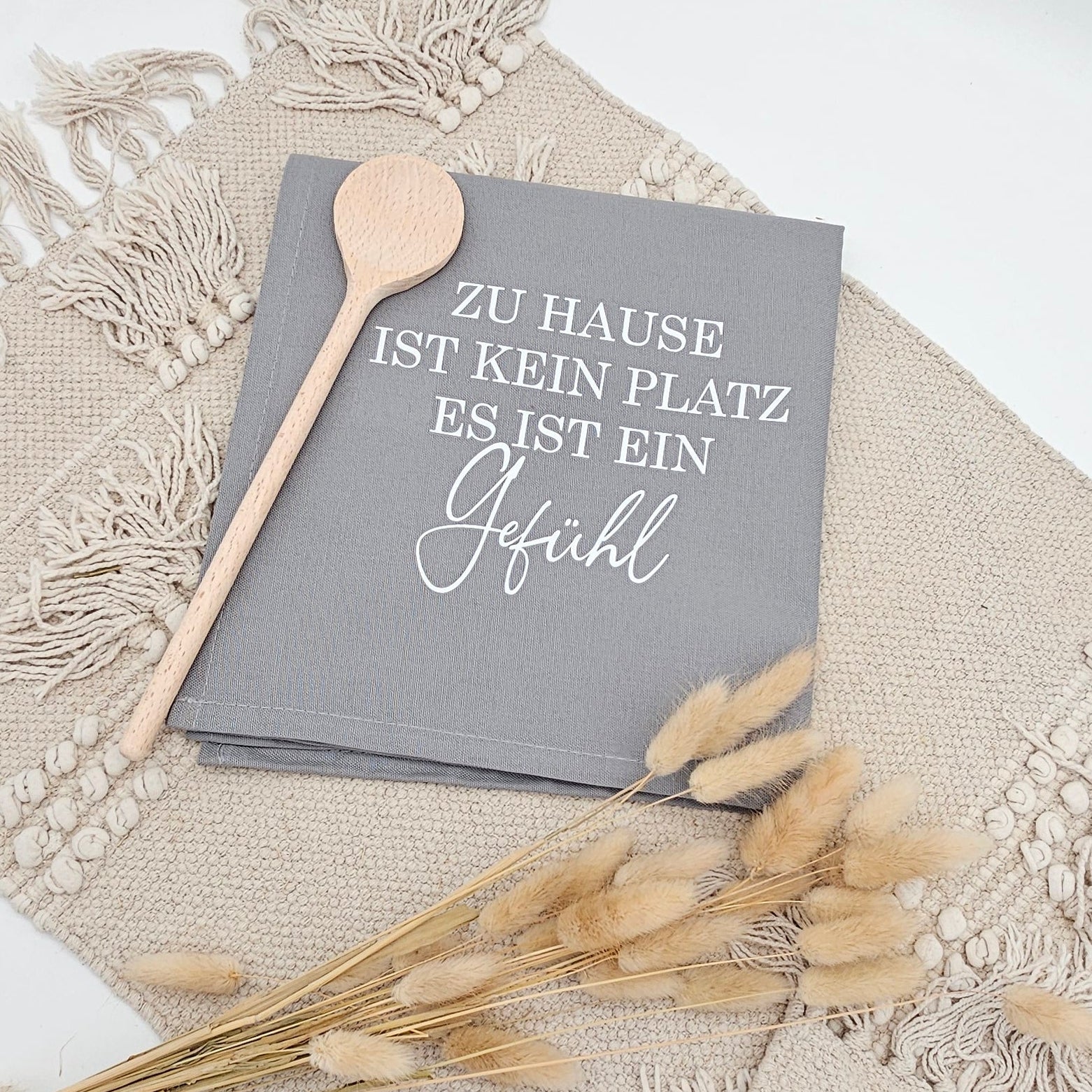 Graues Baumwoll-Geschirrtuch mit weißem Aufdruck „Zu Hause ist kein Platz, es ist ein Gefühl“ neben einem Holzlöffel und Trockenblumen auf einer hellen Boho Unterlage.