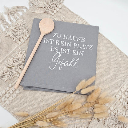 Graues Baumwoll-Geschirrtuch mit weißem Aufdruck „Zu Hause ist kein Platz, es ist ein Gefühl“ neben einem Holzlöffel und Trockenblumen auf einer hellen Boho Unterlage.