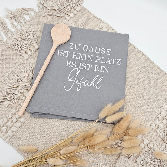Graues Baumwoll-Geschirrtuch mit weißem Aufdruck „Zu Hause ist kein Platz, es ist ein Gefühl“ neben einem Holzlöffel und Trockenblumen auf einer hellen Boho Unterlage.