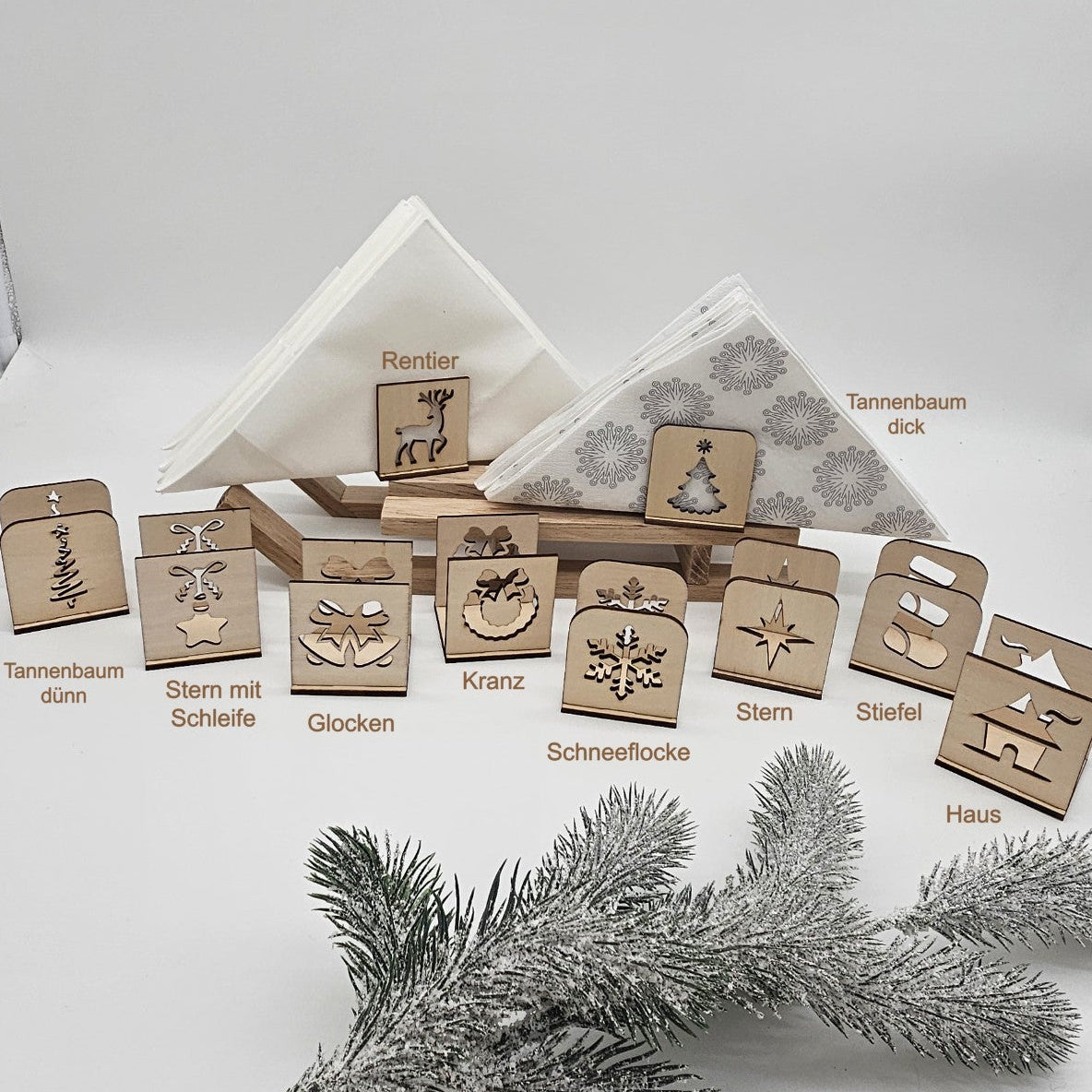 Set aus handgefertigten Holz-Serviettenhaltern mit verschiedenen Weihnachtsmotiven – Tannenbaum, Stern, Geschenk, Kranz und Rentier – auf beiger Tischdecke mit Servietten und Tannenzweig arrangiert.