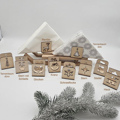 Set aus handgefertigten Holz-Serviettenhaltern mit verschiedenen Weihnachtsmotiven – Tannenbaum, Stern, Geschenk, Kranz und Rentier – auf beiger Tischdecke mit Servietten und Tannenzweig arrangiert.