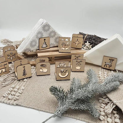 Set aus handgefertigten Holz-Serviettenhaltern mit verschiedenen Weihnachtsmotiven wie Tannenbaum, Stern, Kranz, Rentier und Schneeflocke – arrangiert auf beiger Tischdecke mit Servietten und Tannenzweig im Vordergrund.