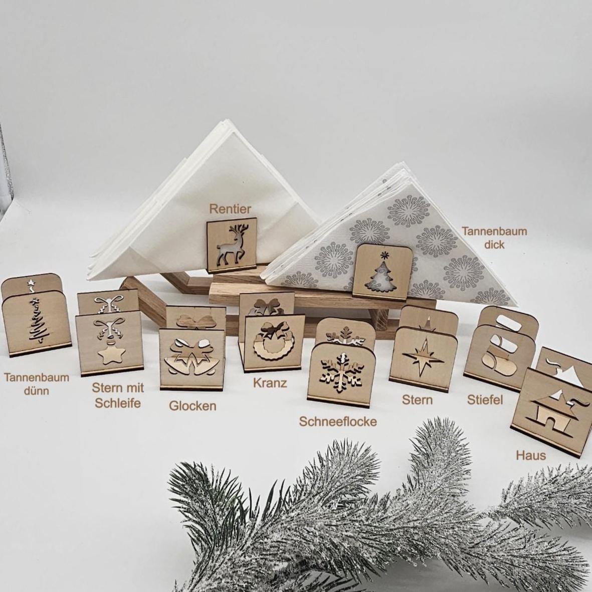 Alle Motive der handgefertigten Holz-Serviettenhalter im Überblick: Tannenbaum, Stern mit Schleife, Glocken, Kranz, Schneeflocke, Rentier, Stern, Stiefel und Haus – präsentiert mit Servietten und Tannenzweig auf hellem Untergrund.