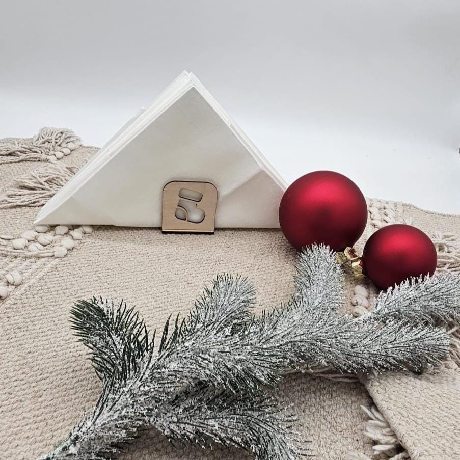 Weihnachtlicher Holz-Serviettenhalter mit Stiefelmotiv, kombiniert mit weißen Servietten, roten Christbaumkugeln und Tannenzweig auf beiger Tischdecke – natürliche Weihnachtsdekoration.