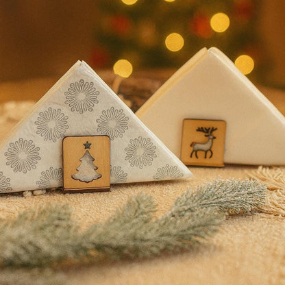 Zwei handgefertigte Holz-Serviettenhalter mit Weihnachtsmotiven – Tannenbaum und Rentier – dekorativ platziert vor warmem, goldenem Hintergrund mit verschwommenen Lichtern und Tannenzweig.