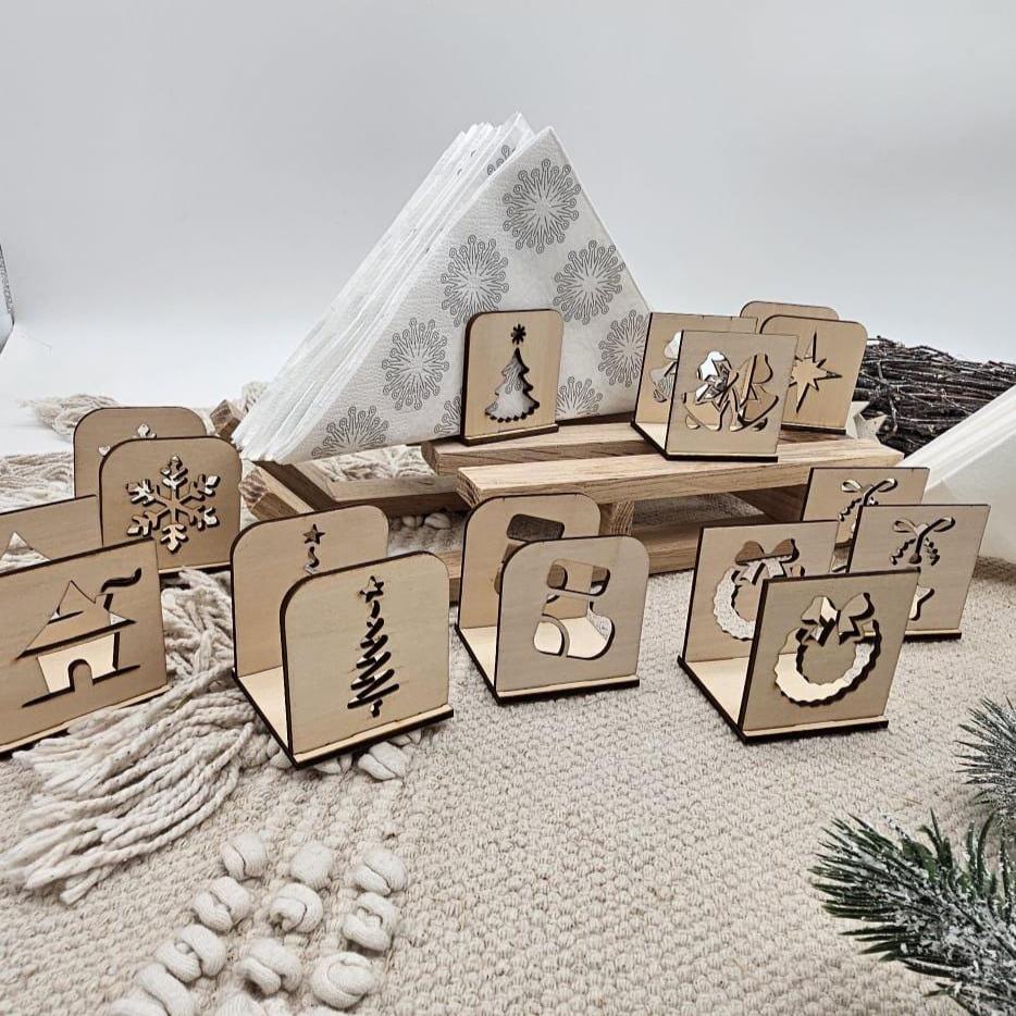 Handgefertigte Holz-Serviettenhalter mit verschiedenen Weihnachtsmotiven – Tannenbaum, Stern, Schneeflocke, Stiefel, Kranz und Haus – auf beiger Tischdecke mit Servietten und Tannenzweig dekorativ angeordnet.