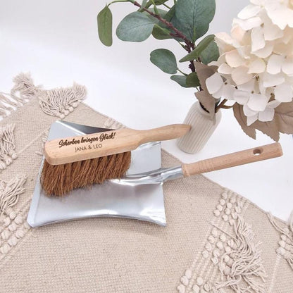 Holzhandfeger mit Gravur 'Scherben bringen Glück! Jana & Leo' auf einer Metallschaufel, umgeben von Boho-Dekoration und Blumen.