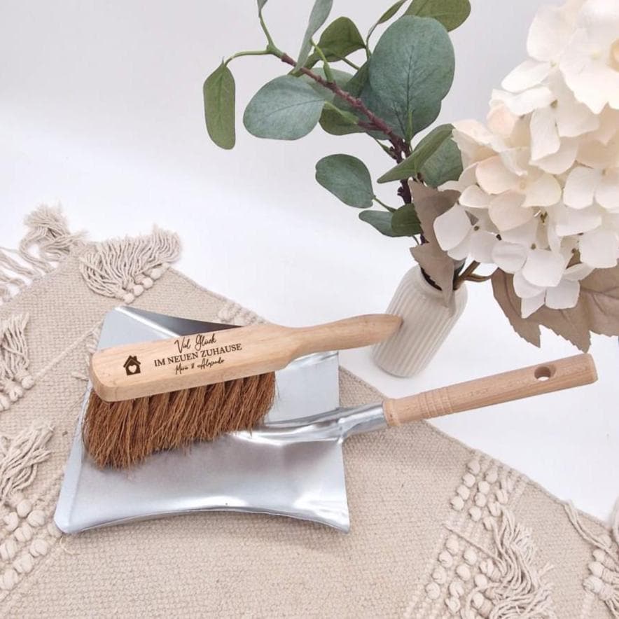 Holzhandfeger mit Gravur 'Viel Glück im neuen Zuhause - Mara & Alexander' und einem kleinen Haus-Symbol auf einer Metallschaufel, dekoriert mit hellen Blumen.