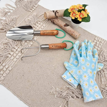 Personalisiertes Garten-Set aus Edelstahl mit Holzgriffen. Gravur 'Gartenkönigin' und 'Sophia' auf Schaufel und Kralle, liegend neben blumigen Gartenhandschuhen.