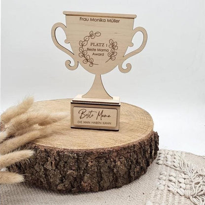 Ein hellbrauner Holz-Pokal mit der Gravur 'Beste Mama Award' und dem Namen 'Frau Monika Müller'. Die Trophäe steht auf einer Baumscheibe neben getrockneten Gräsern