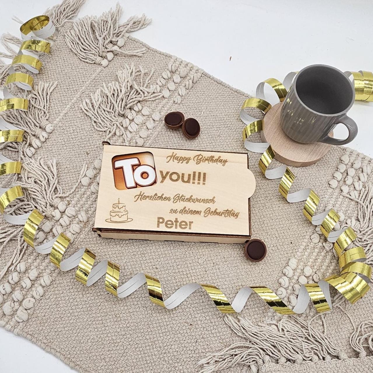 Eine rechteckige Geschenkbox aus hellem Holz mit Schiebedeckel. Der Deckel hat ein ausgeschnittenes 'To'-Sichtfenster und eine Gravur mit dem Namen 'Peter' sowie Glückwünschen zum Geburtstag. Die Box liegt auf einer beigen Stoffmatte mit goldenen Luftschlangen.
