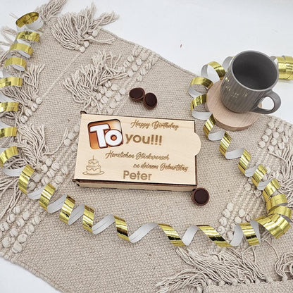 Eine rechteckige Geschenkbox aus hellem Holz mit Schiebedeckel. Der Deckel hat ein ausgeschnittenes 'To'-Sichtfenster und eine Gravur mit dem Namen 'Peter' sowie Glückwünschen zum Geburtstag. Die Box liegt auf einer beigen Stoffmatte mit goldenen Luftschlangen.