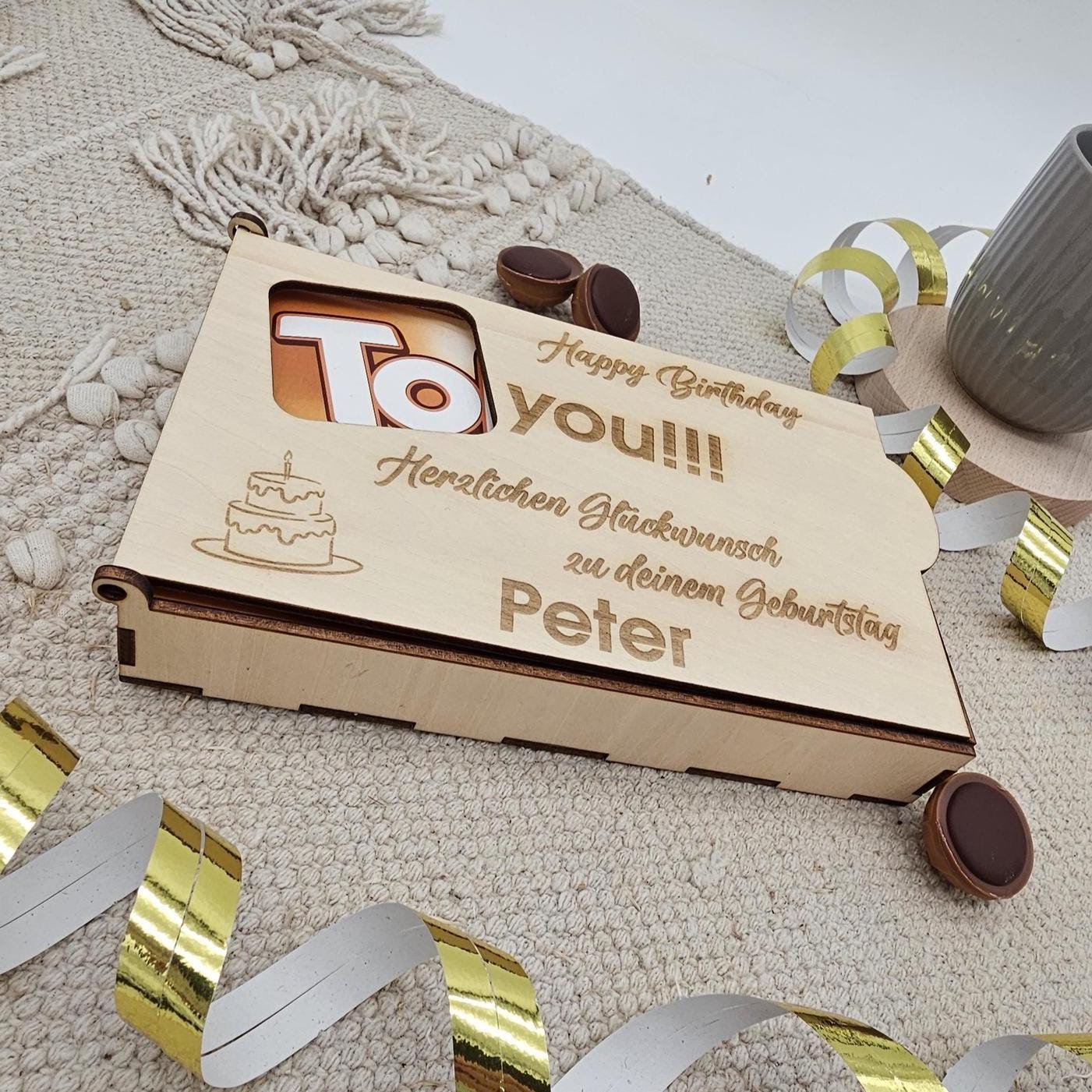 Nahaufnahme einer lasergravierten Holzoberfläche. Zu sehen ist eine kleine Geburtstagstorte und der Text 'Happy Birthday to you!!! Herzlichen Glückwunsch zu deinem Geburtstag Peter' in sauberer, dunkler Schrift.