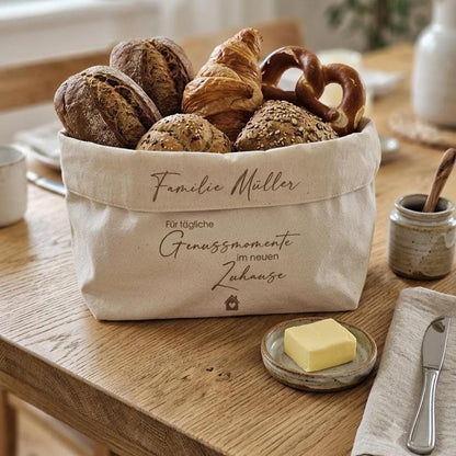 Ein beigefarbener Stoffkorb aus festem Baumwolle mit der braunen Aufschrift 'Familie Müller – Für tägliche Genussmomente im neuen Zuhause' steht auf einem hellen Holztisch. Der Korb ist reichlich gefüllt mit verschiedenen frischen Brötchen, einem Croissant und einer Laugenbrezel. Im Vordergrund liegt ein kleines Keramiktellerchen mit einem Stück Butter, daneben eine Leinenserviette mit einem Messer. Der Hintergrund zeigt ein gemütlich und hell belichtetes Esszimmer mit einem Fenster.