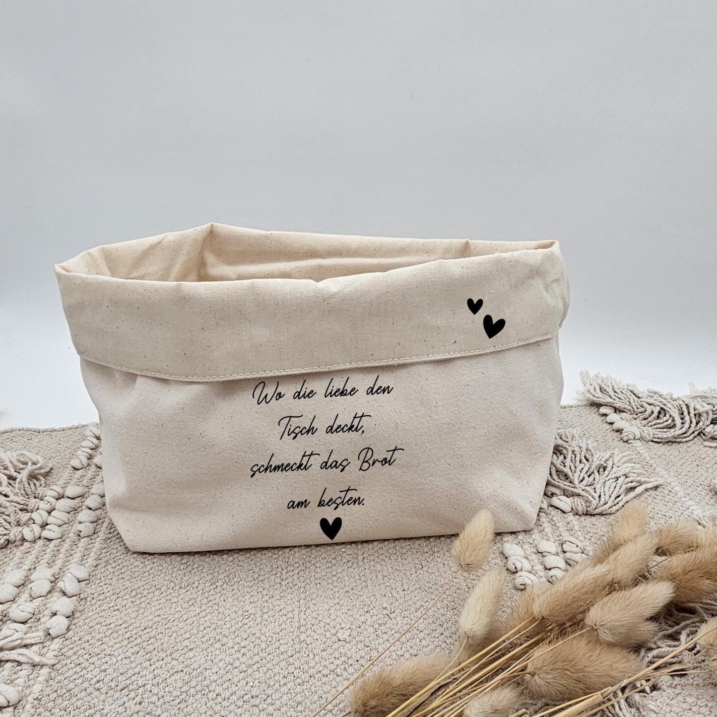 Ein beigefarbener Stoffkorb aus Canvas mit der Aufschrift 'Wo die Liebe den Tisch deckt, schmeckt das Brot am besten' und kleinen schwarzen Herzen auf einem Boho-Teppich. Im Vordergrund liegen getrocknete Pampasgras-Stängel.