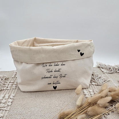 Ein beigefarbener Stoffkorb aus Canvas mit der Aufschrift 'Wo die Liebe den Tisch deckt, schmeckt das Brot am besten' und kleinen schwarzen Herzen auf einem Boho-Teppich. Im Vordergrund liegen getrocknete Pampasgras-Stängel.