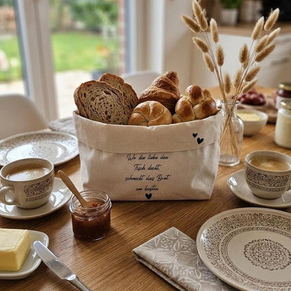 Ein Brotkorb aus Stoff mit einer Backwaren-Auswahl steht auf einem Holztisch, umgeben von einem gedeckten Frühstücksset. In der Mitte des Korbs ist der Text "Wo die Liebe den Tisch deckt, schmeckt das Brot am besten." und zwei Herzen gestickt. Ein Marmeladenglas, Butter, Kaffeetassen und Pampasgras runden das Bild ab. Der Hintergrund ist unscharf.