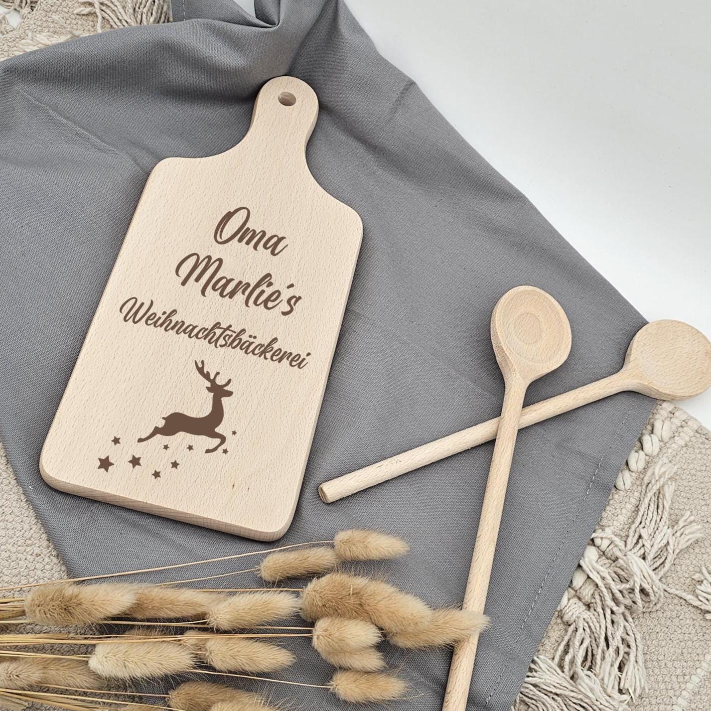 Ein kleines Holzschneidebrett mit Griff liegt auf einem grauen Stofftuch und einem beigen Boho-Teppich. Das Brett ist graviert mit 'Oma Marlie's Weihnachtsbäckerei', einem Rentier und kleinen Sternen. Daneben gekreuzt liegen zwei Holzkochlöffel und ein Bund getrocknete Lagurus-Gräser.