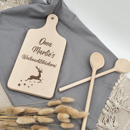 Ein kleines Holzschneidebrett mit Griff liegt auf einem grauen Stofftuch und einem beigen Boho-Teppich. Das Brett ist graviert mit 'Oma Marlie's Weihnachtsbäckerei', einem Rentier und kleinen Sternen. Daneben gekreuzt liegen zwei Holzkochlöffel und ein Bund getrocknete Lagurus-Gräser.