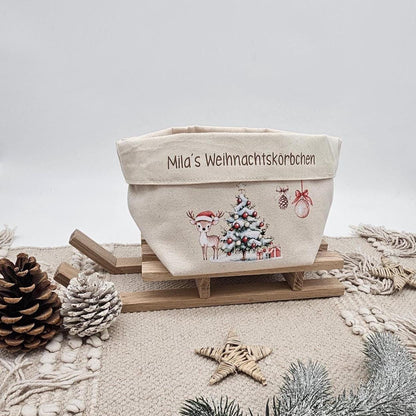 Ein beigefarbener Stoffkorb aus Canvas mit der Aufschrift 'Mila's Weihnachtskörbchen' auf dem Umschlag. Darunter ist ein Motiv mit einem Rehkitz mit Weihnachtsmütze, einem geschmückten Tannenbaum und Christbaumkugeln gedruckt. Der Korb steht auf einem kleinen Holzschlitten auf einem beigen Boho-Teppich. Im Vordergrund liegen Tannenzapfen, ein Strohstiern und ein verschneiter Tannenzweig.