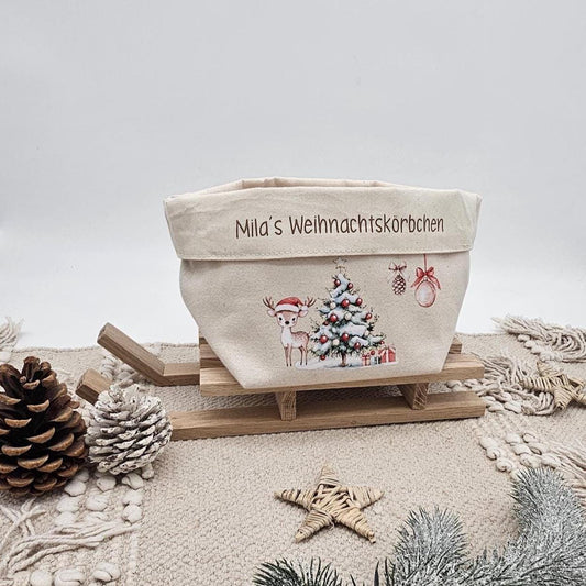 Ein beigefarbener Stoffkorb aus Canvas mit der Aufschrift 'Mila's Weihnachtskörbchen' auf dem Umschlag. Darunter ist ein Motiv mit einem Rehkitz mit Weihnachtsmütze, einem geschmückten Tannenbaum und Christbaumkugeln gedruckt. Der Korb steht auf einem kleinen Holzschlitten auf einem beigen Boho-Teppich. Im Vordergrund liegen Tannenzapfen, ein Strohstiern und ein verschneiter Tannenzweig.