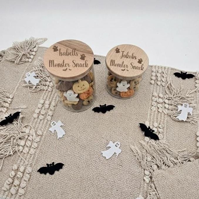 Zwei Vorratsgläser aus Glas mit graviertem Holzdeckel „Isabells Monster Snack“ und „Jakobs Monster Snack“ auf beigem Untergrund mit kleiner Halloween-Deko.