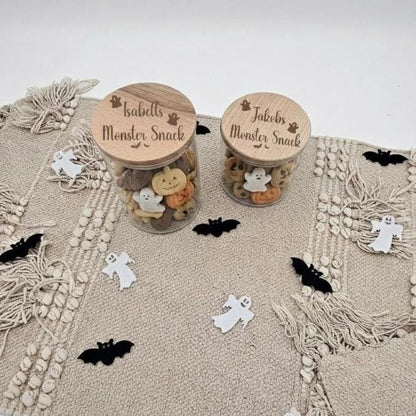 Zwei Vorratsgläser aus Glas mit graviertem Holzdeckel „Isabells Monster Snack“ und „Jakobs Monster Snack“ auf beigem Untergrund mit kleiner Halloween-Deko.