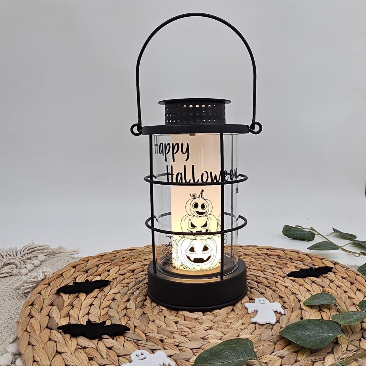 Schwarze Metall-Laterne mit Aufschrift „Happy Halloween“, niedlichem Kürbis-Turm Motiv und warm leuchtender LED-Kerze im Inneren auf Untersetzer.