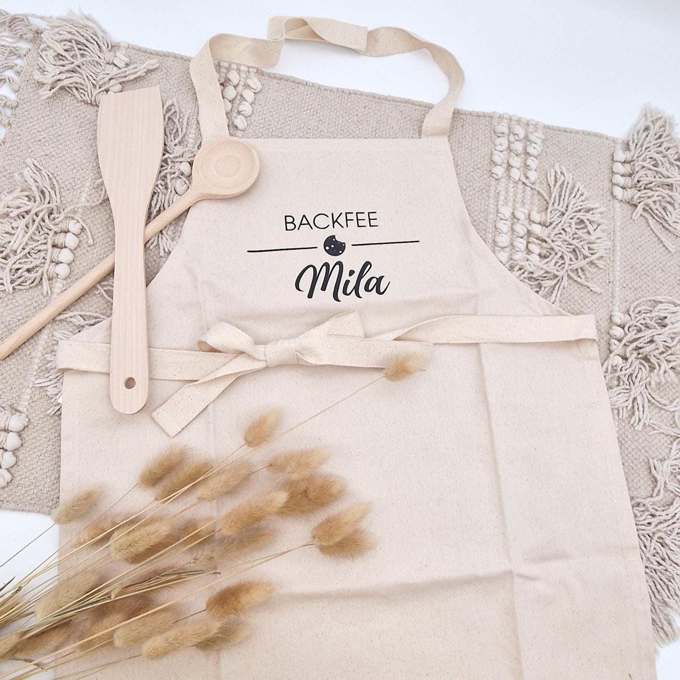 Personalisierte Kinderschürze aus beigefarbener Baumwolle mit Aufschrift „BACKFEE Mila“ und Keks-Motiv, kombiniert mit Holzkochlöffeln im Boho-Stil.