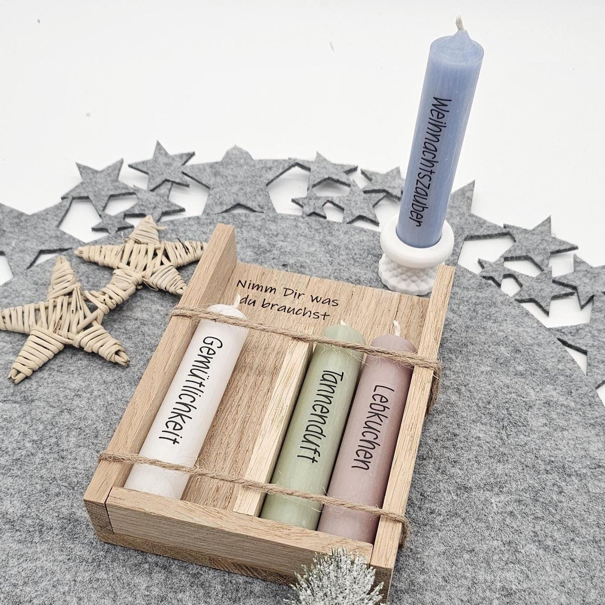Dekoriertes Weihnachtsgeschenkset: Blaue Stabkerze mit Aufschrift ‚Weihnachtszauber‘ im weißen Kerzenhalter stehend, daneben Holzbox ‚Nimm Dir was du brauchst‘ mit weiteren Motivkerzen auf grauer Filzunterlage.
