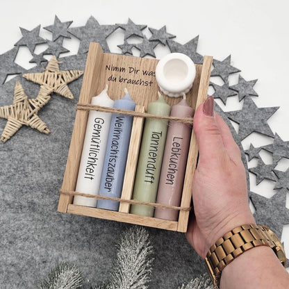 Eine Hand hält ein Weihnachtsgeschenkset in Holzbox mit Aufschrift ‚Nimm Dir was du brauchst‘: Vier bunte Stabkerzen mit weihnachtlichen Sprüchen und weißer Kerzenständer
