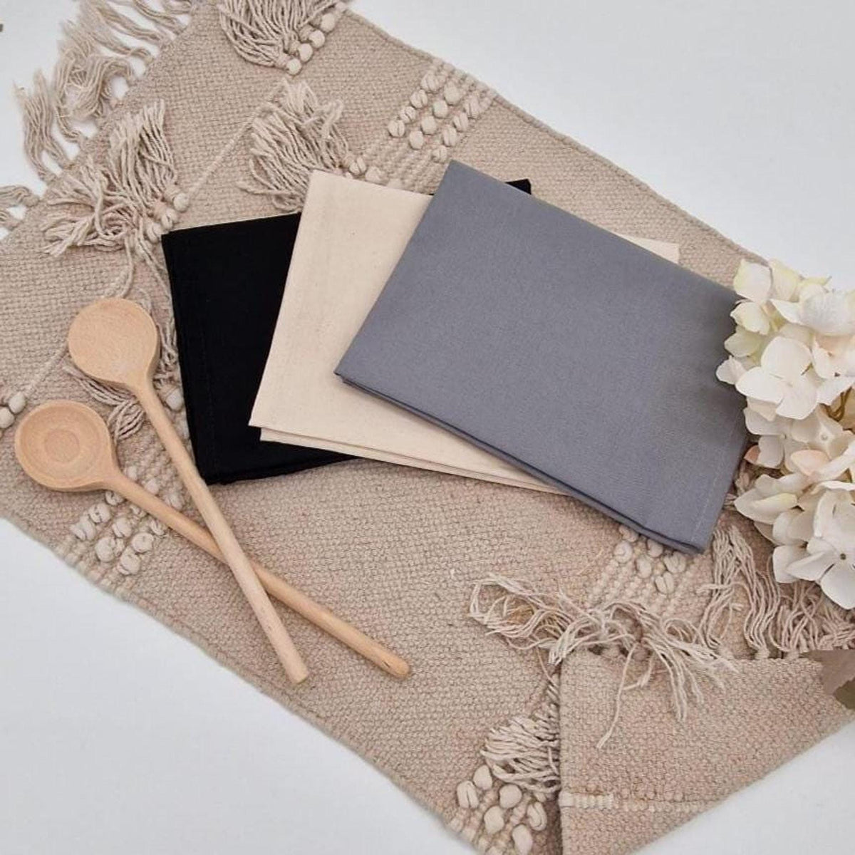 Farb-Auswahl personalisierbarer Geschirrtücher: Gefaltete Stofftücher in Grau, Beige und Schwarz mit Kochlöffeln aus Holz auf Boho-Tischset mit Fransen.