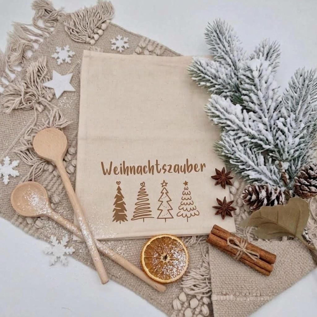 Eine weihnachtliche Flachbild-Aufnahme zeigt einen Leinenbeutel mit dem Aufdruck „Weihnachtszauber“ und vier verschiedenen Tannenbaum-Grafiken. Der Beutel liegt auf einer Fransendecke und ist umgeben von tief verschneiten Tannenzweigen, Kiefernzapfen, Holz löffeln, Sternanis, einer getrockneten Orangenscheibe und Zimtstangen. Weiße Filzschneeflocken tragen zur adventlichen Atmosphäre bei. Der Hintergrund ist weiß.