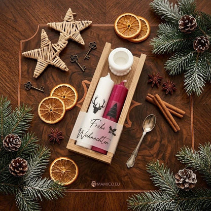Festliches Weihnachts-Flatlay mit Kerzenset in einer Holzleiste auf dunklem Holzuntergrund. Die Holzbox enthält eine weiße Kerze mit Hirsch-Motiv, eine rote Kerze mit Tannenbaum-Motiv und einen weißen Kerzenhalter. Umwickelt mit Banderole „Frohe Weihnachten“. Dekoriert mit Weidensternen, getrockneten Orangenscheiben, Zimtstangen, Sternanis und verschneiten Tannenzweigen. MAMICO.EU Logo unten.