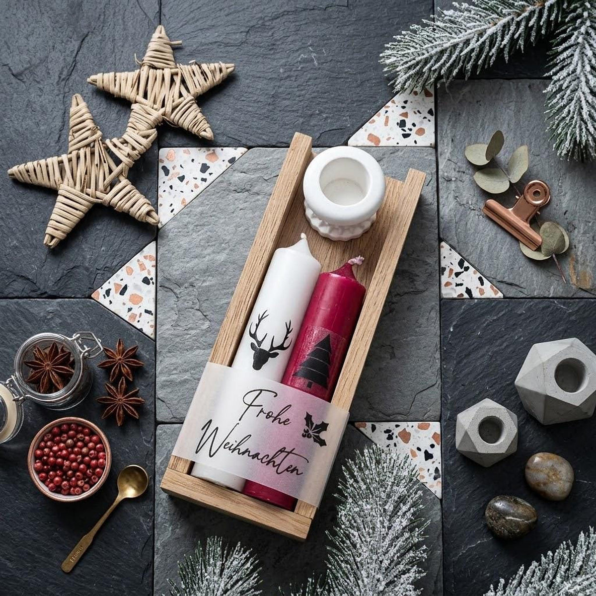 Modernes Weihnachts-Flatlay auf Schiefer- und Terrazzofliesen. Eine Holzleiste zeigt ein Kerzenset mit weißer Hirsch-Kerze, roter Tannenbaum-Kerze und weißem Halter, umwickelt mit der Banderole „Frohe Weihnachten“. Umgeben von Weidensternen, Sternanis, rosa Beeren, verschneiten Tannenzweigen und Beton-Deko.