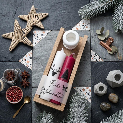 Modernes Weihnachts-Flatlay auf Schiefer- und Terrazzofliesen. Eine Holzleiste zeigt ein Kerzenset mit weißer Hirsch-Kerze, roter Tannenbaum-Kerze und weißem Halter, umwickelt mit der Banderole „Frohe Weihnachten“. Umgeben von Weidensternen, Sternanis, rosa Beeren, verschneiten Tannenzweigen und Beton-Deko.