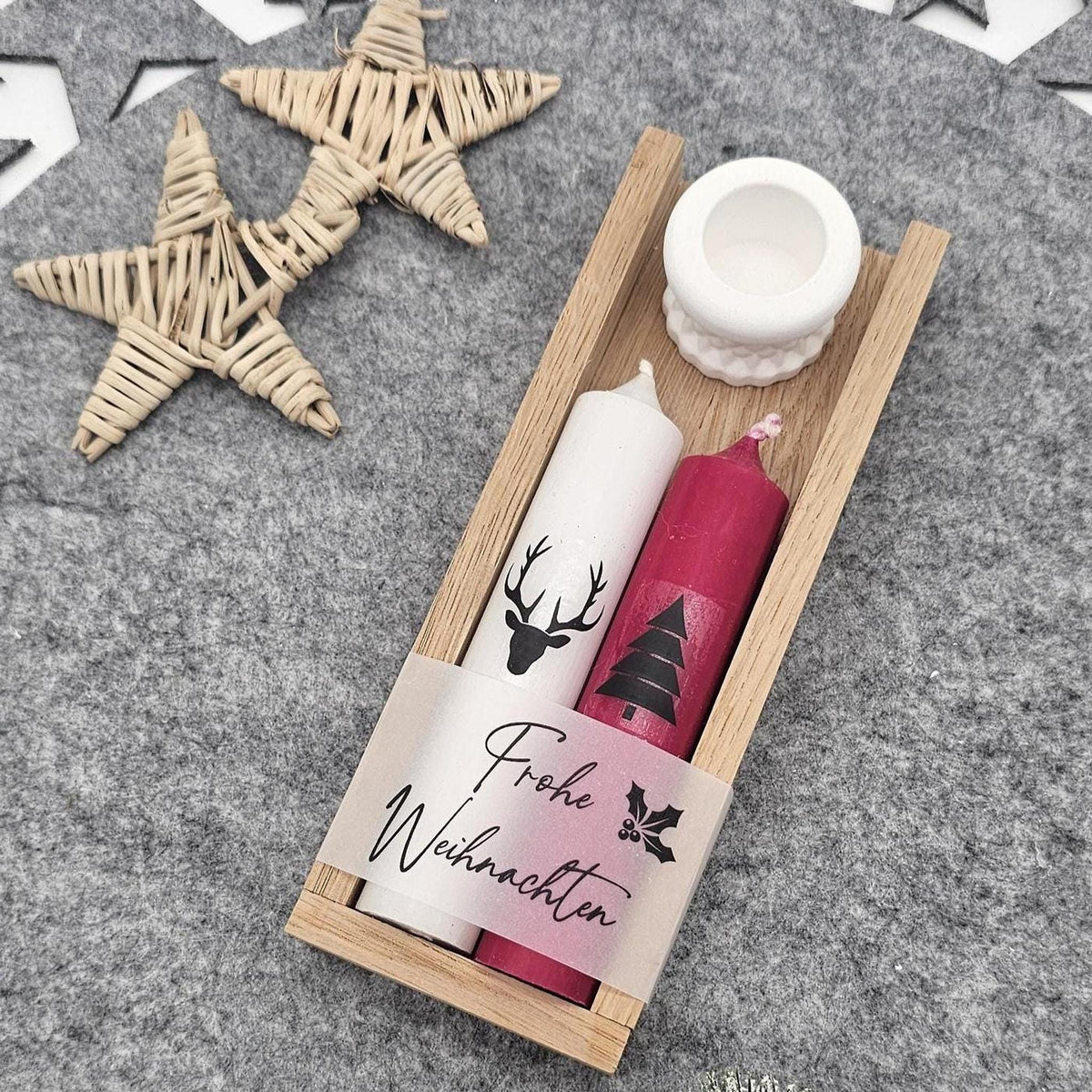 Weihnachtsgeschenkset in flacher Holzbox aus der Vogelperspektive: Weiße Stabkerze mit Hirsch-Motiv, rote Kerze mit Tannenbaum, ‚Frohe Weihnachten‘-Banderole und weißer Kerzenständer auf grauer Filzunterlage.