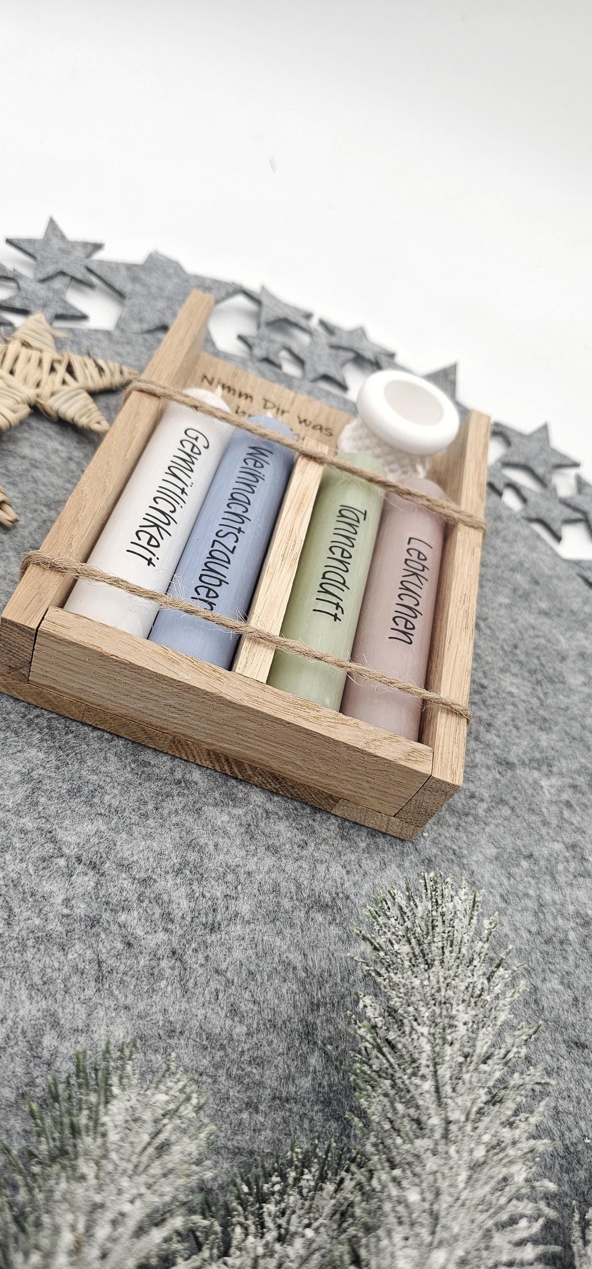 Nahaufnahme von unten: Weihnachtsgeschenkset in Holzbox mit vier bunten Motivkerzen (Gemütlichkeit, Weihnachtszauber, Tannenduft, Lebkuchen) und weißem Kerzenständer auf grauer Filzunterlage.