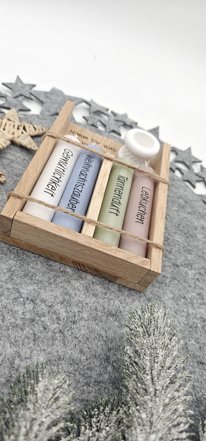 Nahaufnahme von unten: Weihnachtsgeschenkset in Holzbox mit vier bunten Motivkerzen (Gemütlichkeit, Weihnachtszauber, Tannenduft, Lebkuchen) und weißem Kerzenständer auf grauer Filzunterlage.