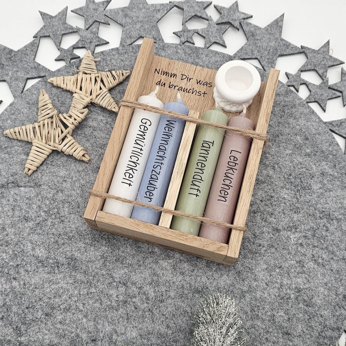 Weihnachtsgeschenkset in Holzbox mit Spruch ‚Nimm Dir was du brauchst‘: Vier bunte Stabkerzen mit Aufschrift Gemütlichkeit, Weihnachtszauber, Tannenduft und Lebkuchen sowie weißem Kerzenhalter auf grauer Filzunterlage.