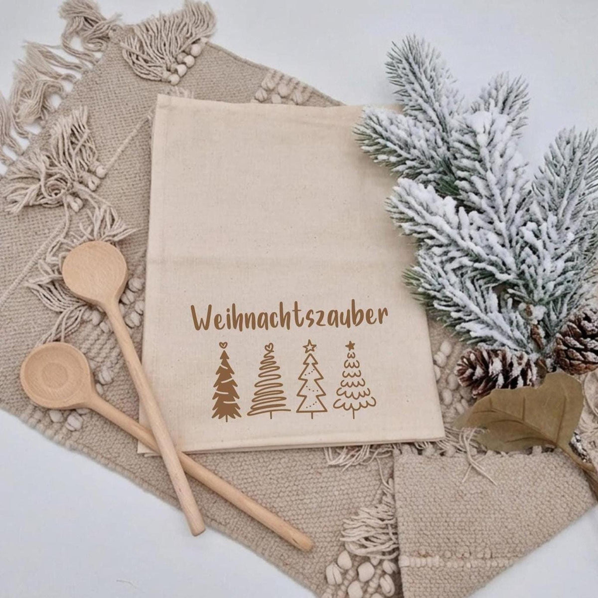 Weihnachtsgeschenk: Beige Baumwollschürze mit Aufschrift ‚Weihnachtszauber‘ und vier Tannenbaum-Illustrationsmotiven, Kochlöffeln aus Holz und Tannenzweig auf boho Stoffunterlage.
