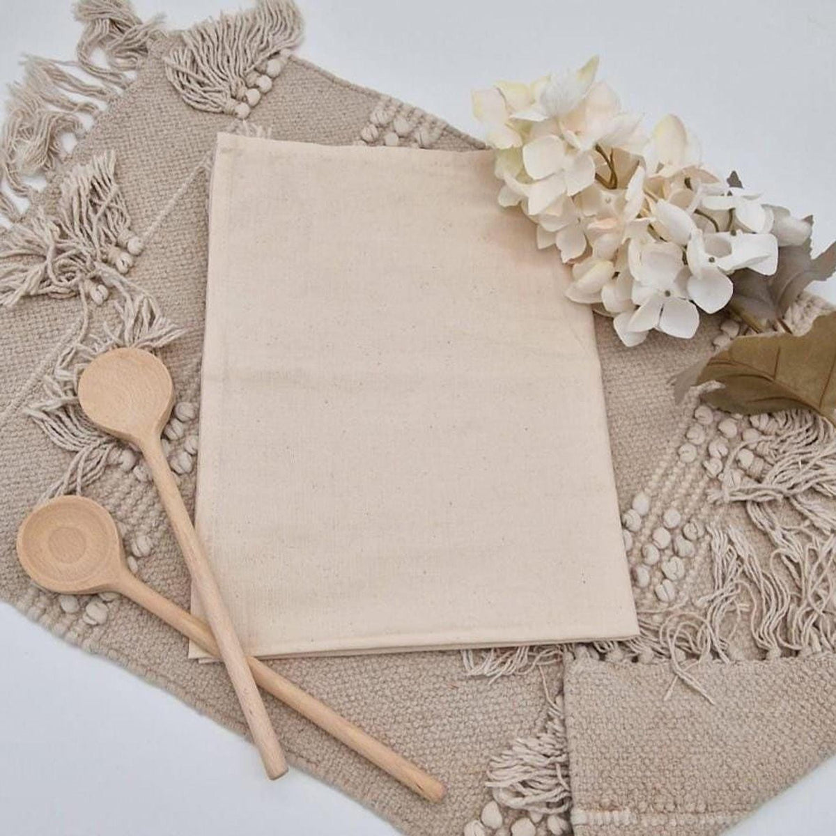 Personalisiertes Geschirrtuch aus beigefarbener Baumwolle ohne Aufdruck, dekoriert mit zwei Holzlöffeln und hellen Hortensienblüten auf einem Boho-Tischset mit Fransen