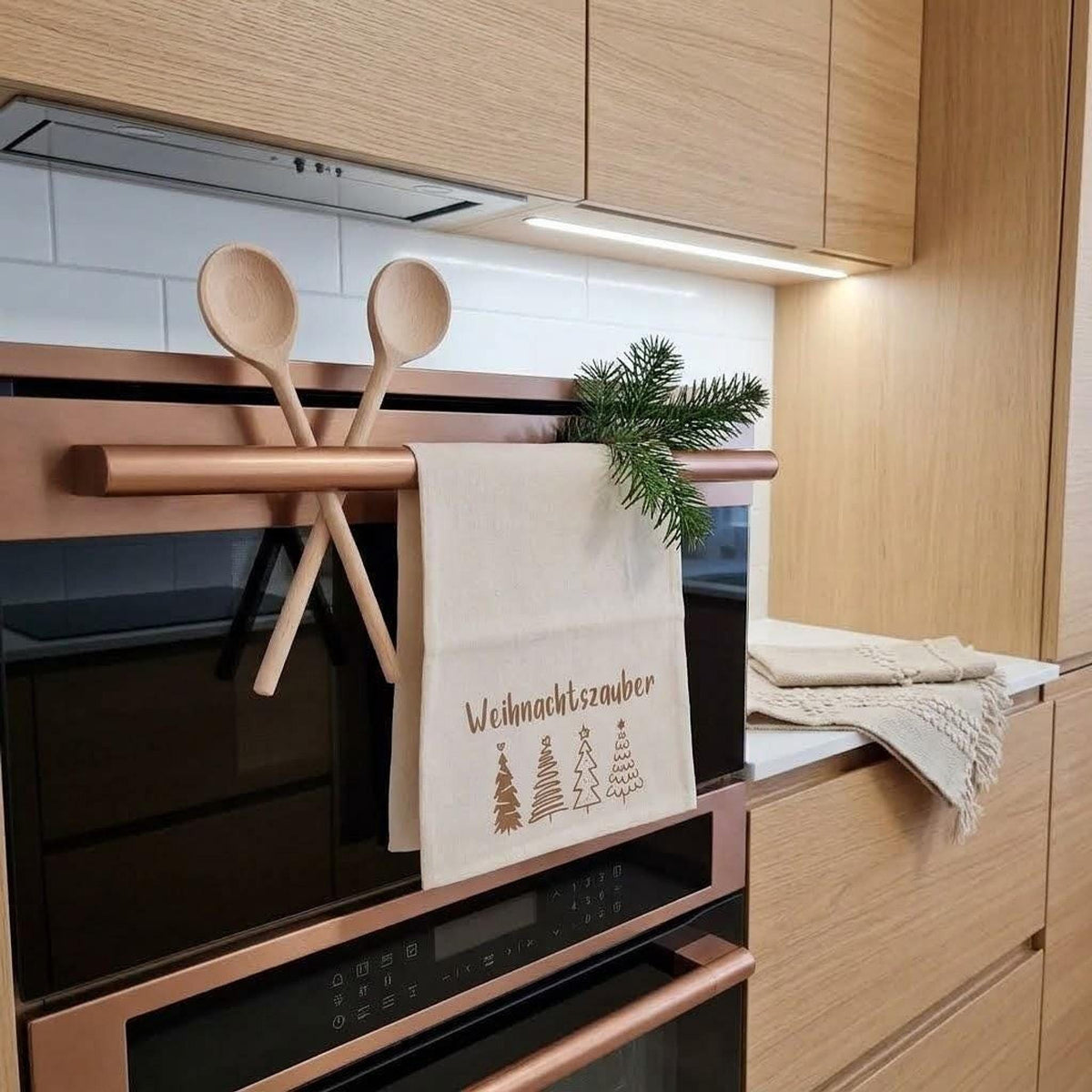 Das Bild zeigt eine moderne Küche mit kupferfarbenem Backofen. Am Griff hängt ein Leinentuch mit der Aufschrift „Weihnachtszauber“ und vier Baumgrafiken. Zwei Kochlöffel sind gekreuzt und ein Tannenzweig steckt im Griff. Auf der Theke liegt eine weitere gemusterte Decke. Die Schränke sind aus Holz.