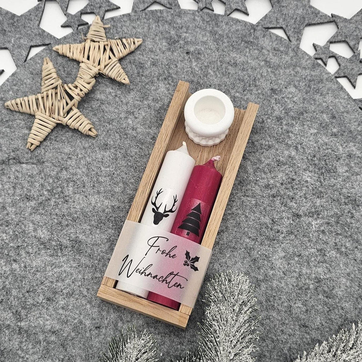 Handgemachtes Weihnachtsgeschenk-Set in Holzbox: Enthält eine weiße Stabkerze mit Hirsch, eine rote Motivkerze mit Tannenbaum und „Frohe Weihnachten“-Banderole sowie einen weißen Kerzenhalter auf weihnachtlich dekoriertem Filz.