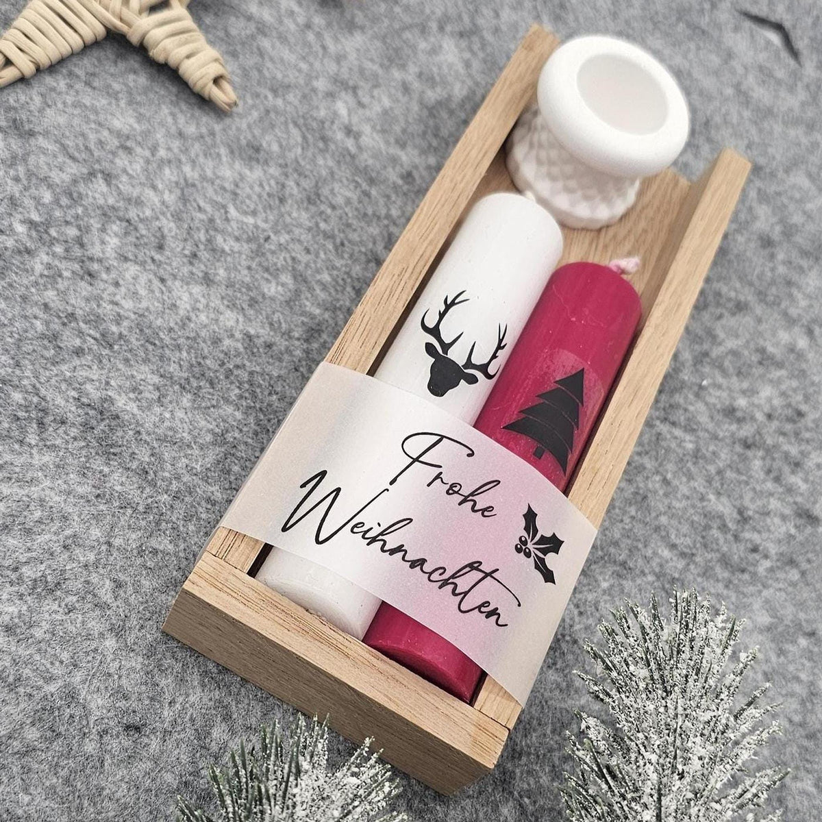 Handgefertigtes Weihnachtsgeschenkset in Holzbox: Weiße Stabkerze mit Hirsch-Motiv, rote Kerze mit Tannenbaum und Banderole ‚Frohe Weihnachten‘ inklusive weißem Kerzenständer auf grauer Filzunterlage, in Nahaufnahme.