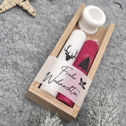 Handgefertigtes Weihnachtsgeschenkset in Holzbox: Weiße Stabkerze mit Hirsch-Motiv, rote Kerze mit Tannenbaum und Banderole ‚Frohe Weihnachten‘ inklusive weißem Kerzenständer auf grauer Filzunterlage, in Nahaufnahme.