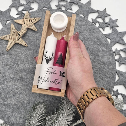 Eine Hand hält ein handgefertigtes Weihnachtsgeschenkset in einer Holzbox: Weiße Stabkerze mit Hirsch, rote Kerze mit Tannenbaum und ‚Frohe Weihnachten‘-Banderole sowie weißer Kerzenhalter.