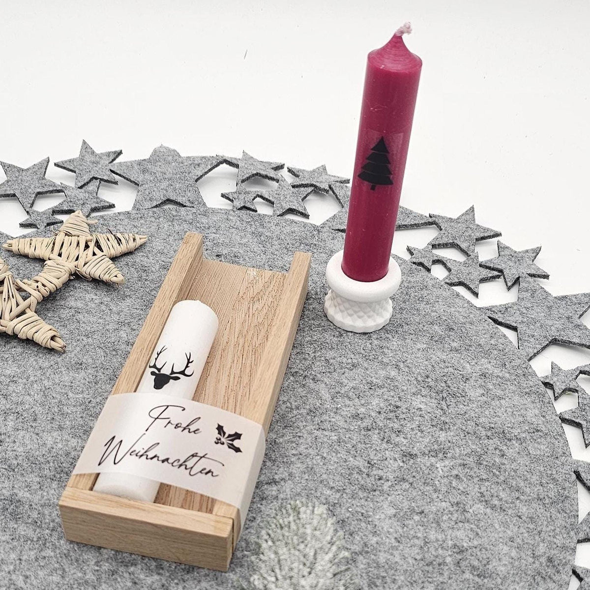 Dekoriertes Weihnachtsgeschenkset: Rote Stabkerze mit Tannenbaum im weißen Kerzenhalter stehend, daneben weiße Hirsch-Kerze in Holzbox mit Banderole ‚Frohe Weihnachten‘ auf grauer Filzunterlage.