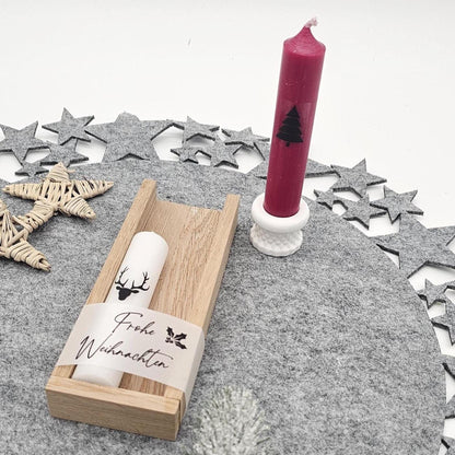 Dekoriertes Weihnachtsgeschenkset: Rote Stabkerze mit Tannenbaum im weißen Kerzenhalter stehend, daneben weiße Hirsch-Kerze in Holzbox mit Banderole ‚Frohe Weihnachten‘ auf grauer Filzunterlage.