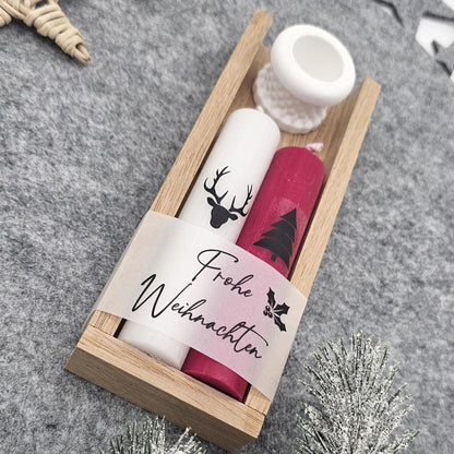 Nahaufnahme eines Weihnachtsgeschenksets in Holzschale: Weiße Stabkerze mit Hirsch, rote Kerze mit Tannenbaum und Banderole ‚Frohe Weihnachten‘ sowie kleiner weißer Kerzenhalter auf grauer Filzunterlage.